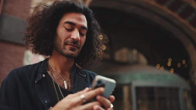 Young attractive italian guy with long curly hair and stubble is using mobile phone at old buildings background. Stylish man with an earring in his ear and lot of chains looking on smartphone