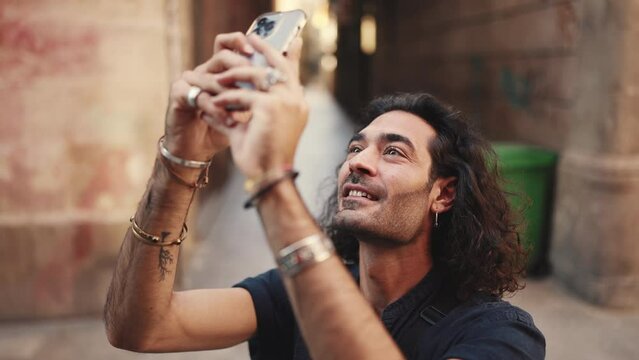 Young attractive italian guy with long curly hair and stubble takes photo of an old building. Stylish man with an earring in his ear and lot of chains photographing sights. Сamera moves around