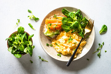 Scrambled eggs, sandwich with cream cheese and salmon and salad leaves at white stone table.