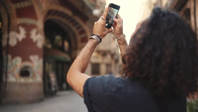 Young attractive italian guy with long curly hair and stubble takes photo of an old building. Stylish man with an earring in his ear and lot of chains photographing sights. Back view