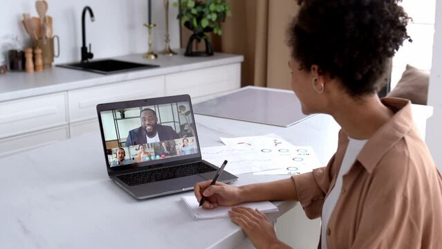 Side view of African American girl to a laptop screen with multiracial people. Female brainstorming with work colleagues, listening to online webinar using laptop and video conference, takes notes