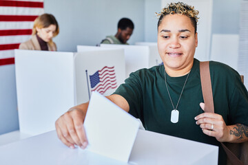 Portrait of modern American woman with tattooes voting and putting ballot in bin on election day,...
