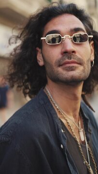 VERTICAL VIDEO: Young italian guy with long curly hair and stubble is walking along crosswalk. Stylish man with sunglasses and lot of chains crosses the road and looks around