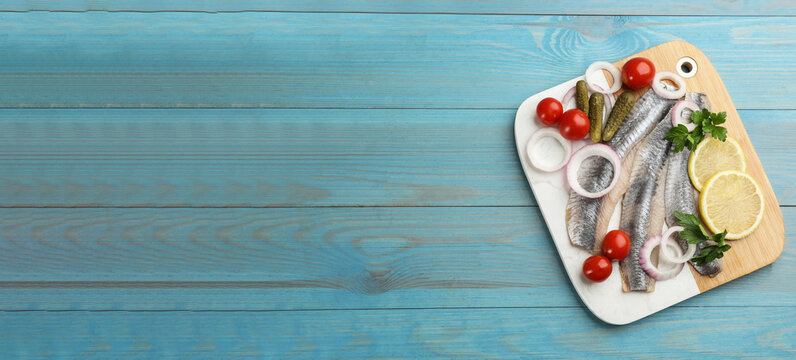 Serving board with salted herring fillets, parsley, onion rings, pickles, cherry tomatoes and lemon on light blue wooden table, top view with space for text. Banner design