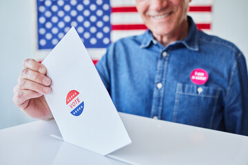 Man putting ballot in voting box, close up with American flag in background, copy space