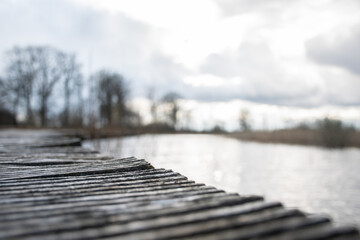 A dreamy picture of a landscape with a close up of an old wooden bridge over a lake with clouds and sunlight and trees in the background