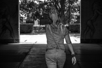 A black and white picture of a caucasian woman with blond hair walking through a dark gate to the light with her hand on her shoulder
