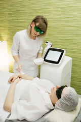 A beautiful girl lies in the office of a beautician in goggles from laser irradiation, the hands of the doctor remove hair on hands with a laser device. Vertical photo