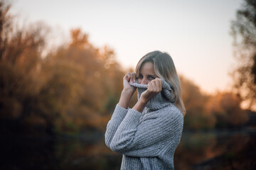 Charming blonde woman standing by river in forest, looking away smiling and pulling sweater collar over face with trees covered with sunset in background. Autumn walk in forest.