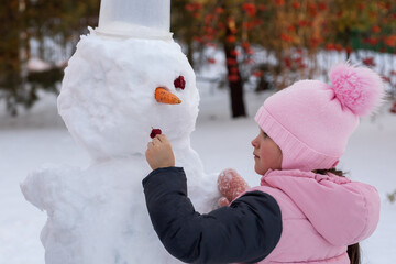 Little female kid making snowman face with help of improvised means in daytime while having walk in...