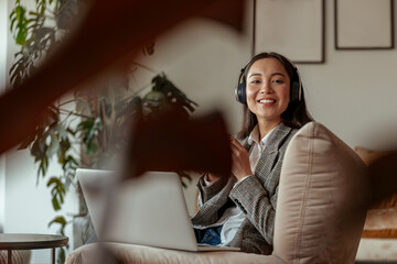 Attractive asian business woman listening music sitting with laptop in office and looking camera