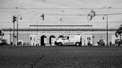 A black and white picture of a street scenery in Vienna Austria. With street lights and driving cars and people walking. In the background big arches to a large palace