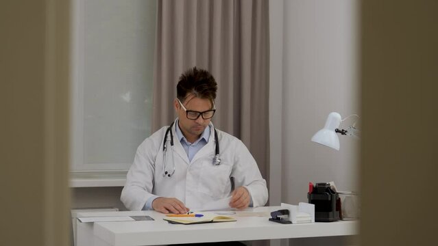 a male doctor in glasses looks at an x-ray, compares the data and writes them down in a notebook view through the gap in the doorway