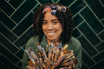 Portrait of young female artist holding dirty brushes and looking at camera and smiling in creative studio - Powered by Adobe