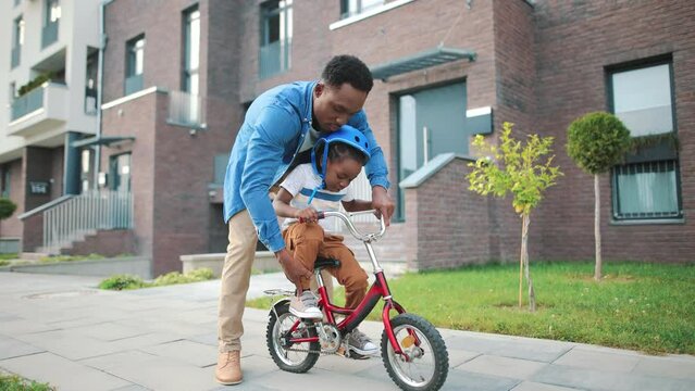 Handsome African American young man parent teaching his small son how to ride a bike outside in neighbourhood area. Little cute boy child in helmet learning to ride a bicycle with dad, happy childhood