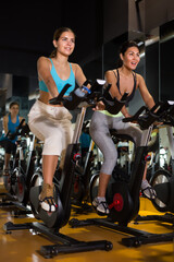 Fototapeta na wymiar Positive girl with her female friend riding exercise bike during cycling class in gym