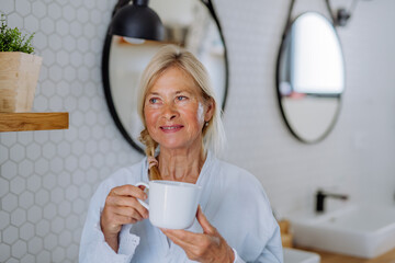 Beautiful senior woman in bathrobe drinking tea in bathroom, relax and wellness concept.