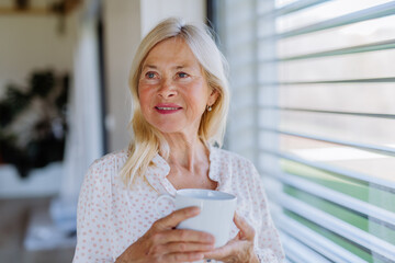 Attractive senior woman drinking tea and standing by window at home.