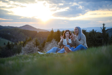 Happy senior grandmother with teenage granddaguhter sitting and whistling with grass in nature on...