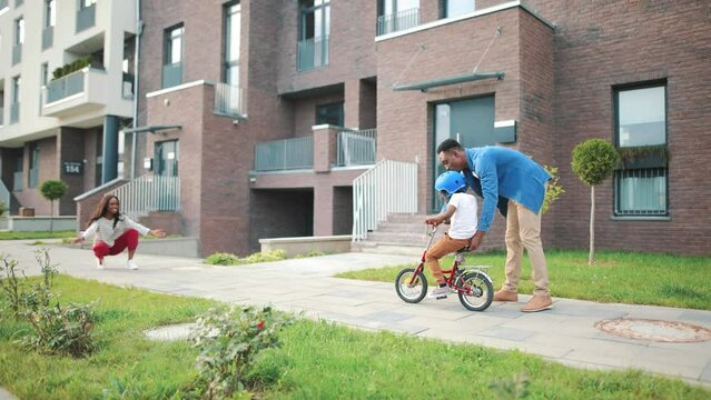 Handsome African American young dad teaching his small son how to ride a bicycle outside in neighbourhood area. Little cute boy child on bike with dad riding toward pretty happy mom, family concept