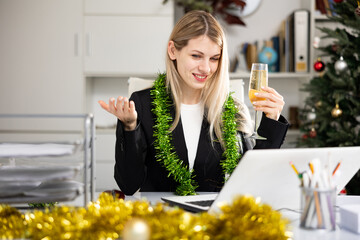 Young woman manager sitting at table in Christmas decorated office with glass of champagne and...