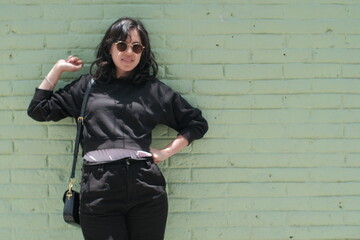 happy young black-haired woman dressed in black sweatshirt and black pants and sunglasses leaning against a mint colored brick wall grabbing her hair and smiling