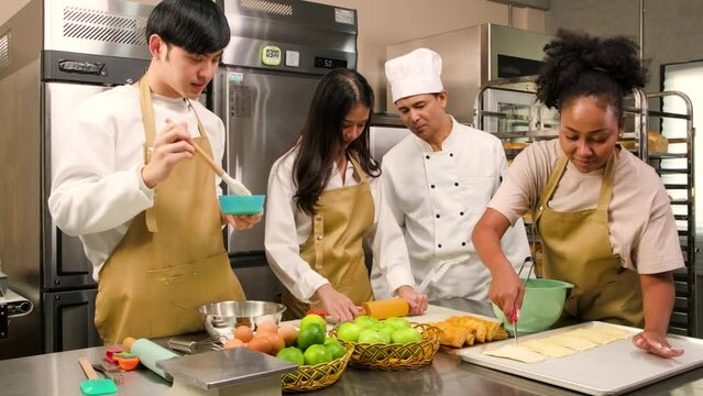 Cuisine course, a senior male chef in cook uniform teaches young cooking class students to prepare ingredients and pastry dough for bakery foods and fruit pies in a restaurant stainless steel kitchen.