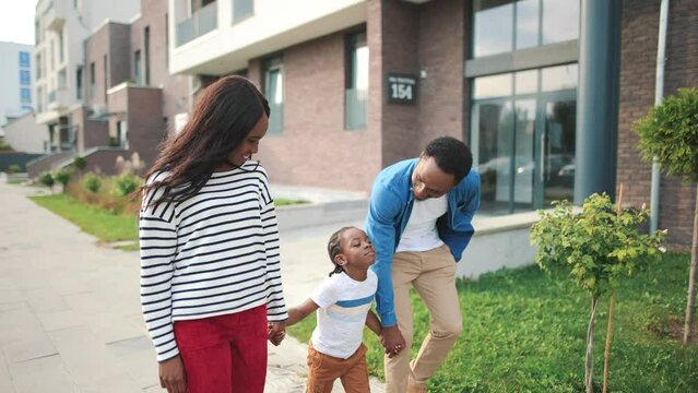 Joyful nice African American young family on a walk outdoors. Happy beautiful mother and handsome father walking with cute small son holding hands and smiling. Family day, child concept