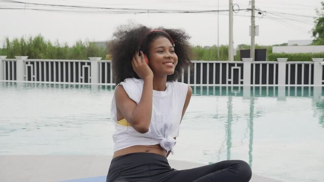 Young African American woman wearing headphones listening music at swimming pool, female wearing earphones while enjoy with happiness at poolside, emotion and feeling, lifestyles concept.