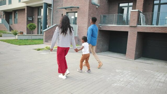 Rear of African American young lovely parents and kid on a walk in the city. Happy little child walking with mom and dad in neighbourhood holding hands. Leisure, family time, childhood concept