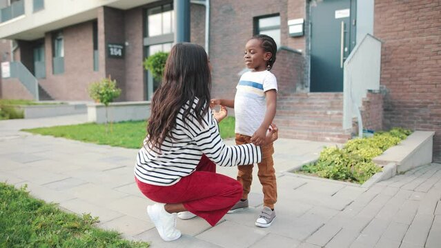 Rear of young beautiful African american woman parent talking with little kid boy and embracing him outdoors in city. Caring mother chatting with kid and gives him a hug on street, family concept