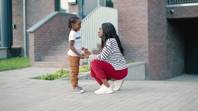 Side view of African American little cute cheerful kid boy speaking with young pretty mom on street and smiling. Caring loving mother chatting with small son outdoor in town. Family concept