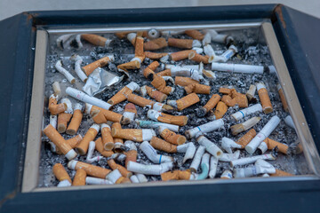 Many of cigarette butts in a big public ash tray.  Stacked cigarette butts.