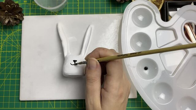 Female artist draws muzzle with brush on clay rabbit. Plastic palette, acrylic paint and rabbit figurine on table. Leisure and crafts in quarantine.
