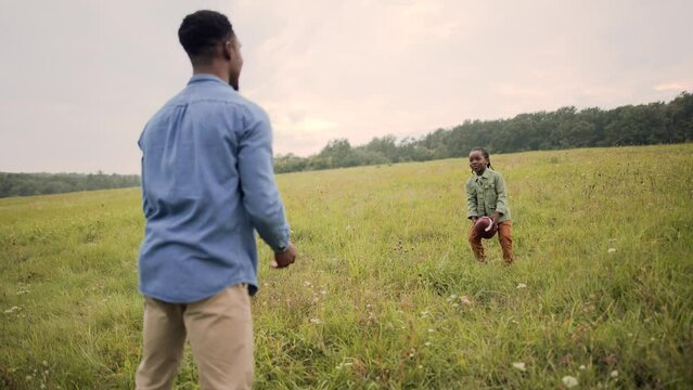 African American caring father playing with little son kid throw and catch with ball standing in field outdoors. Dad teaching child to play american football in meadow. Happy childhood. Family concept