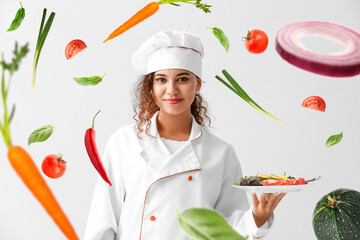 Young African-American female chef with healthy dish and flying vegetables on light background