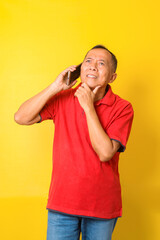 Senior man talking to telephone and thingking, wear red t shirt isolated on yellow background. 