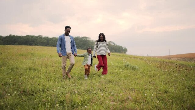 African American young man and woman walking with little kid in meadow holding hands and smiling. Parents with small son in green field outside city spending family day together, parenthood, childhood
