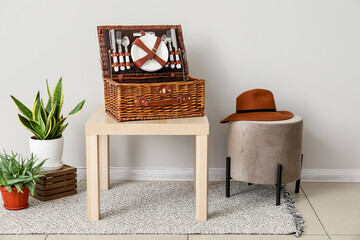 Table with basket for picnic, pouf and houseplants near light wall