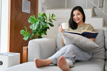 Beautiful young asian woman on sofa having a coffee and reading a book