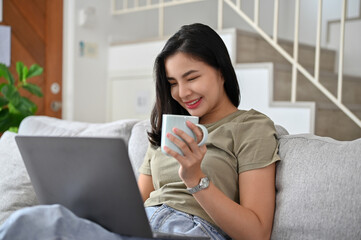 Beautiful young Asian woman having a coffee on the sofa and using laptop computer.