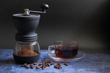 A coffee cup, grinder, and coffee beans are on the table with a black background.
