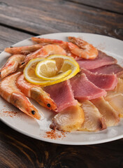 Boiled shrimp and assorted fish fillets on a white plate. A portion of beer snacks on a wooden table close-up. Vertical orientation, copy space, no people