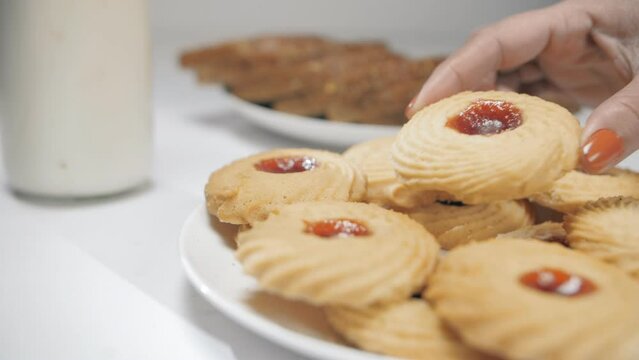 Female's hand keeping a cookie topped with jam on a stack of cookies - unhealthy snack  bad food for heart disease . Closeup shot of mouth-watering biscuits with a glass of milk and bread slices