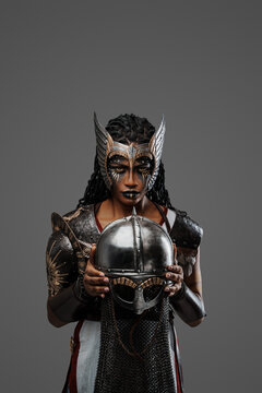 Portrait of black female warrior dressed in ancient armor holding helmet against grey background.