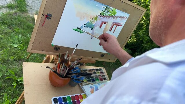 close-up with a brush draw a picture on the picture a house with two windows on the windowsills flowers grow around a lot of greenery grass trees this summer, a brush is held by a male adult hand.