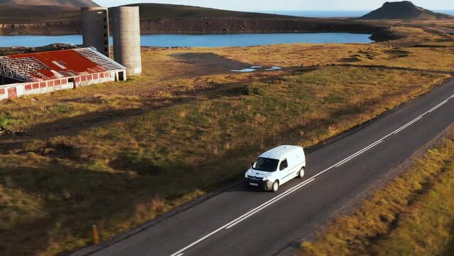 Small white transporter van passing by abandoned farm with old silos, golden hour sunlight, adventure journey