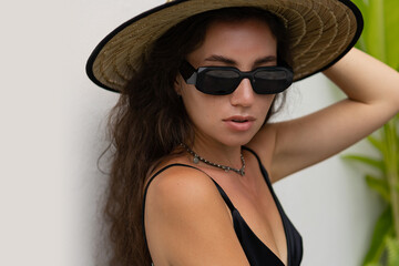 Close up portrait of  beautiful model woman   in trendy sunglases and black summer  outfit  with straw hat posing on modern  villa.