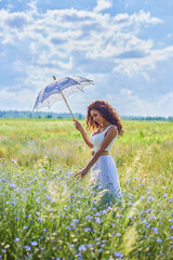 girl in a white summer dress with an umbrella stands among high grass in countryside