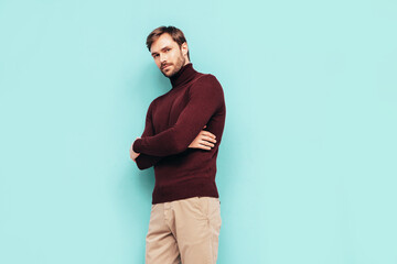 Portrait of handsome confident stylish hipster lambersexual model. Sexy man dressed in turtleneck sweater and trousers. Fashion male isolated on blue wall in studio